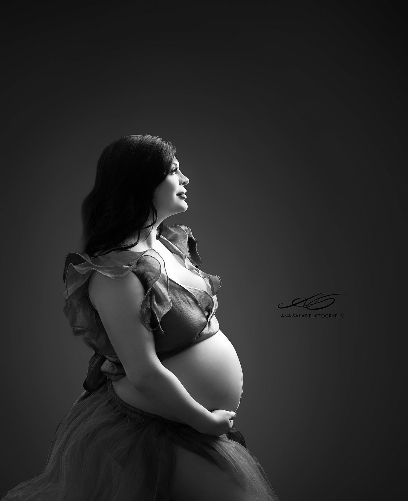 A happy mom to be sits in a spotlight in a studio in an open maternity gown after visiting lucina midwives