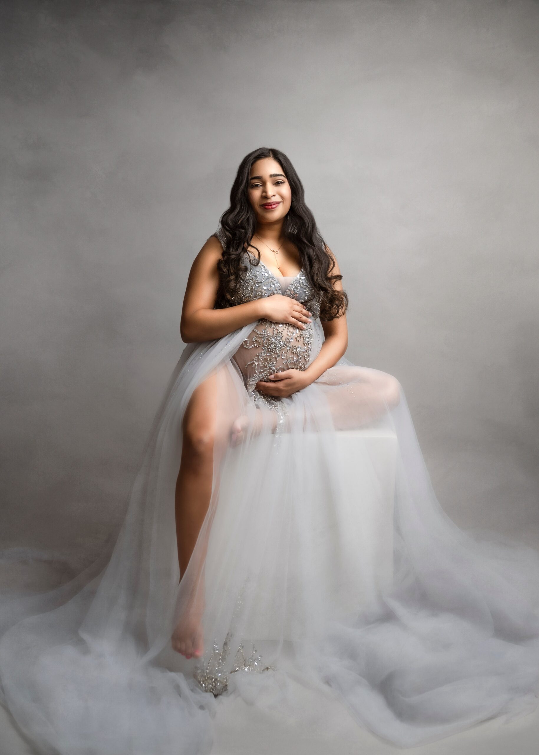A smiling pregnant woman sits on a cube in a long flowing maternity gown