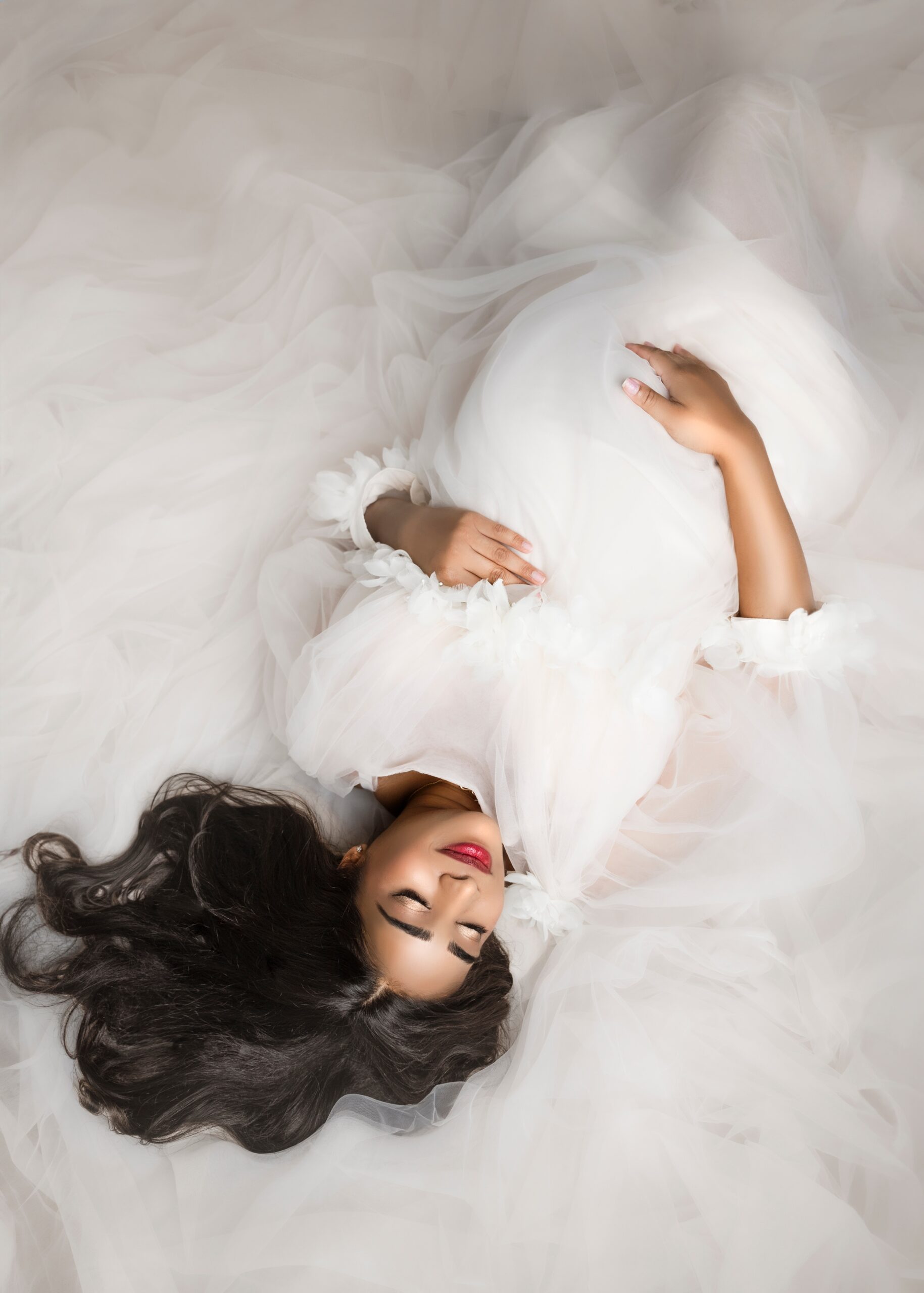 A mom to be lays in the long flowing train of her white maternity gown on the floor of a studio holding her bump with eyes closed after meeting aspen community midwives