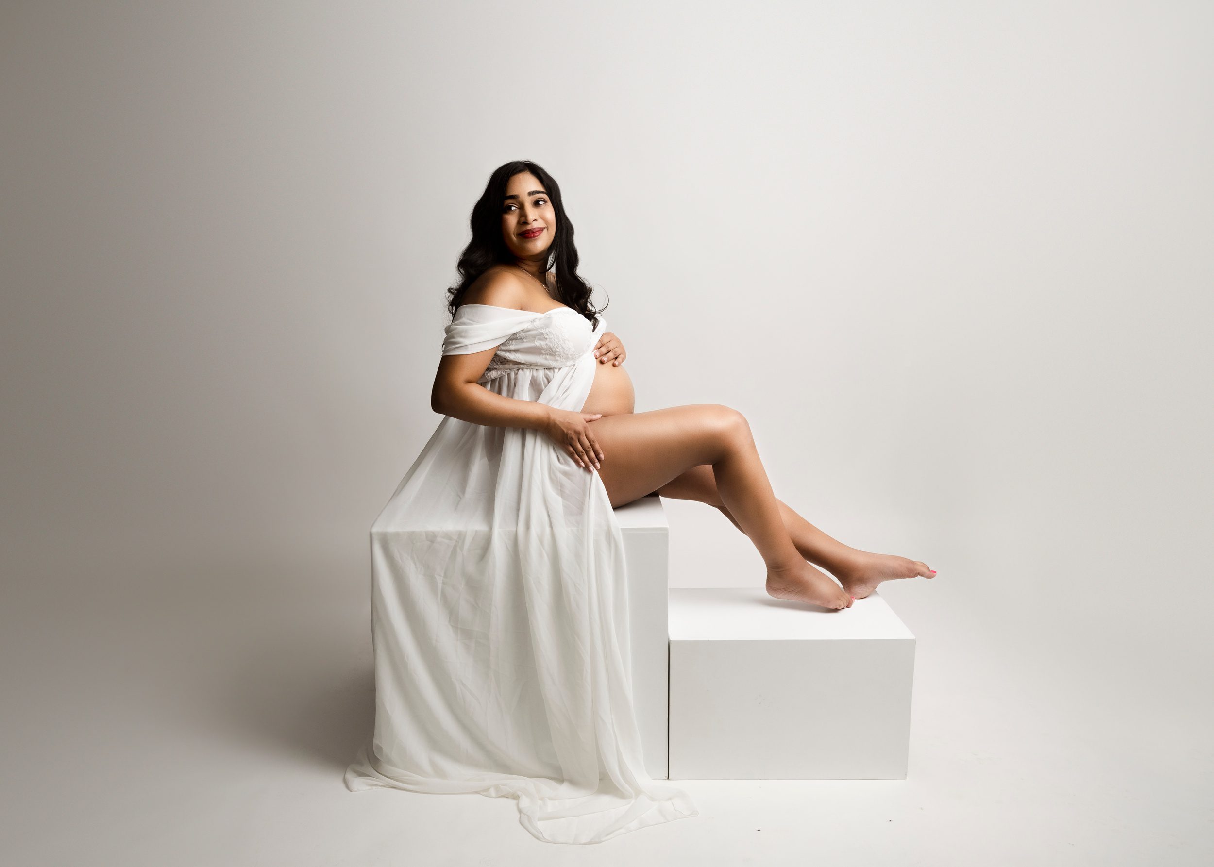 A happy mother to be sits on white cubes in a studio in a long white maternity gown while holding a hand on her bump