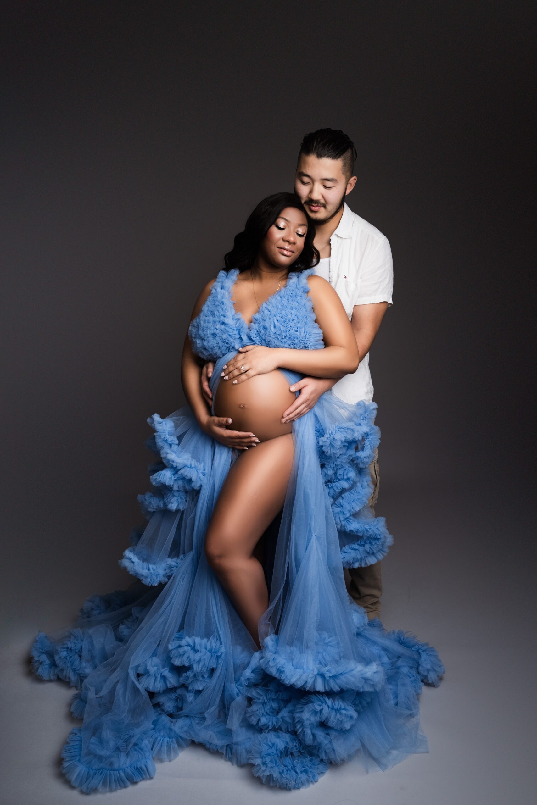 A pregnant woman leans into her husband for snuggles while standing in a studio in a blue gown open in the front
