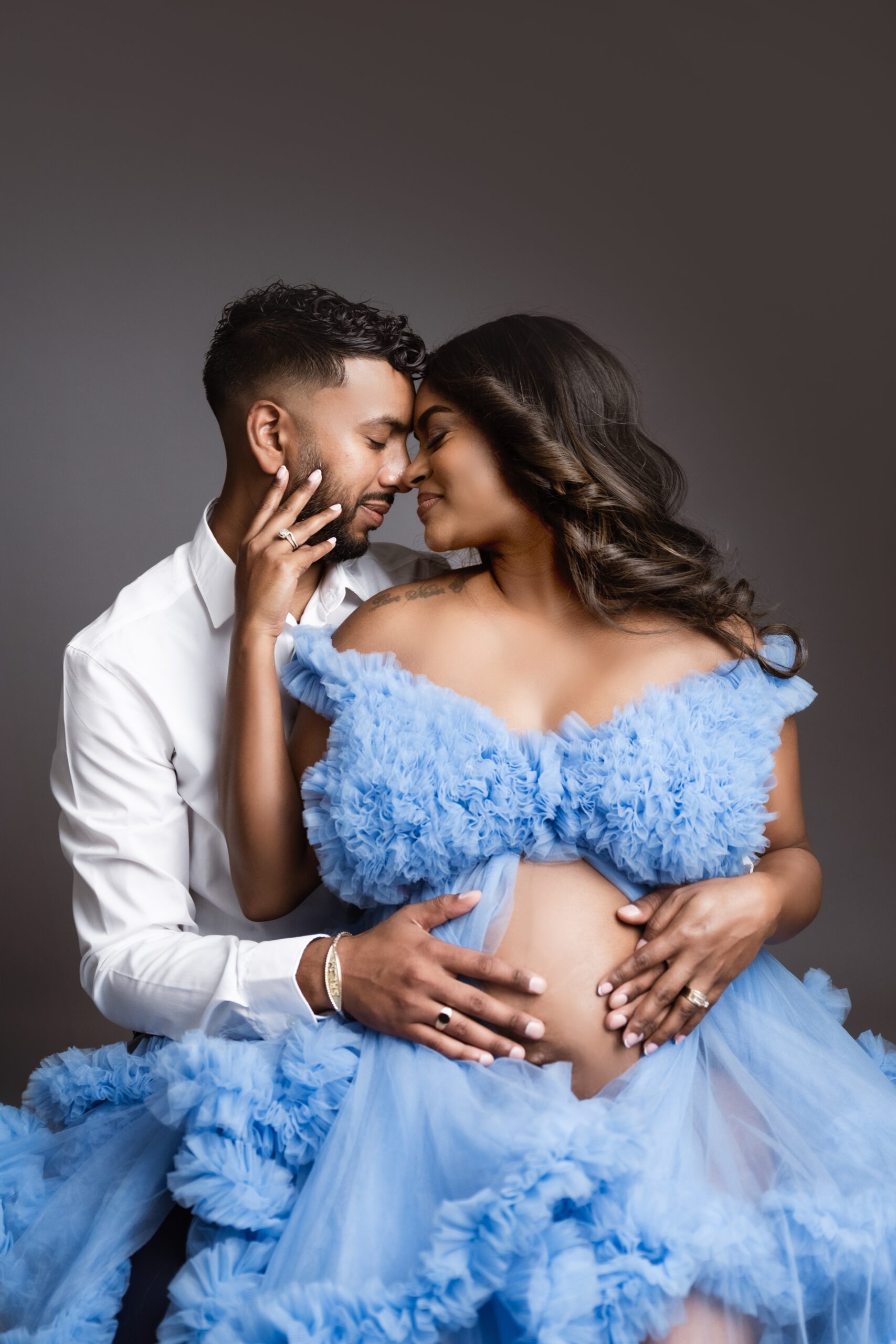 Happy expecting parents snuggle in a studio with hands on bump in an open blue tule maternity gown after visiting full circle birth collective