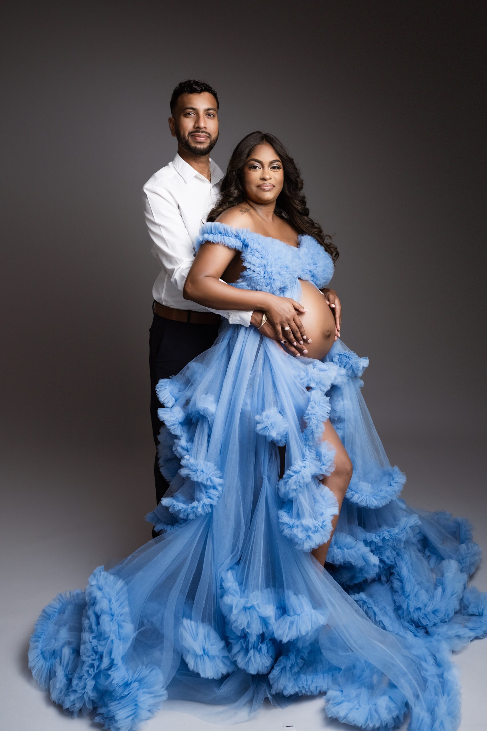 Happy mom to be in a flowing blue tule maternity gown hods the bump with her husband in a studio after visiting full circle birth collective