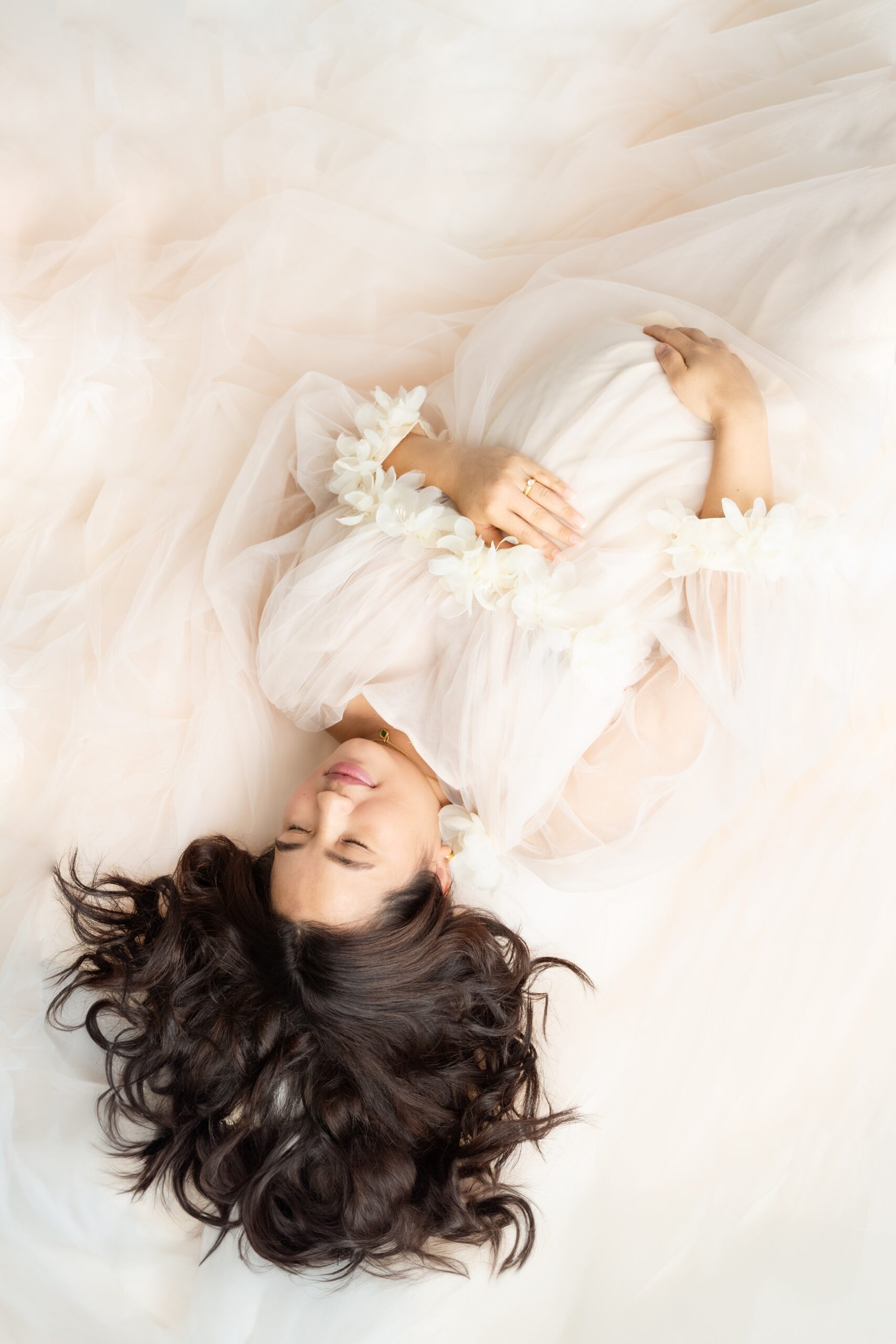 A smiling mom to be lays in a long pink maternity gown with hands on her bump