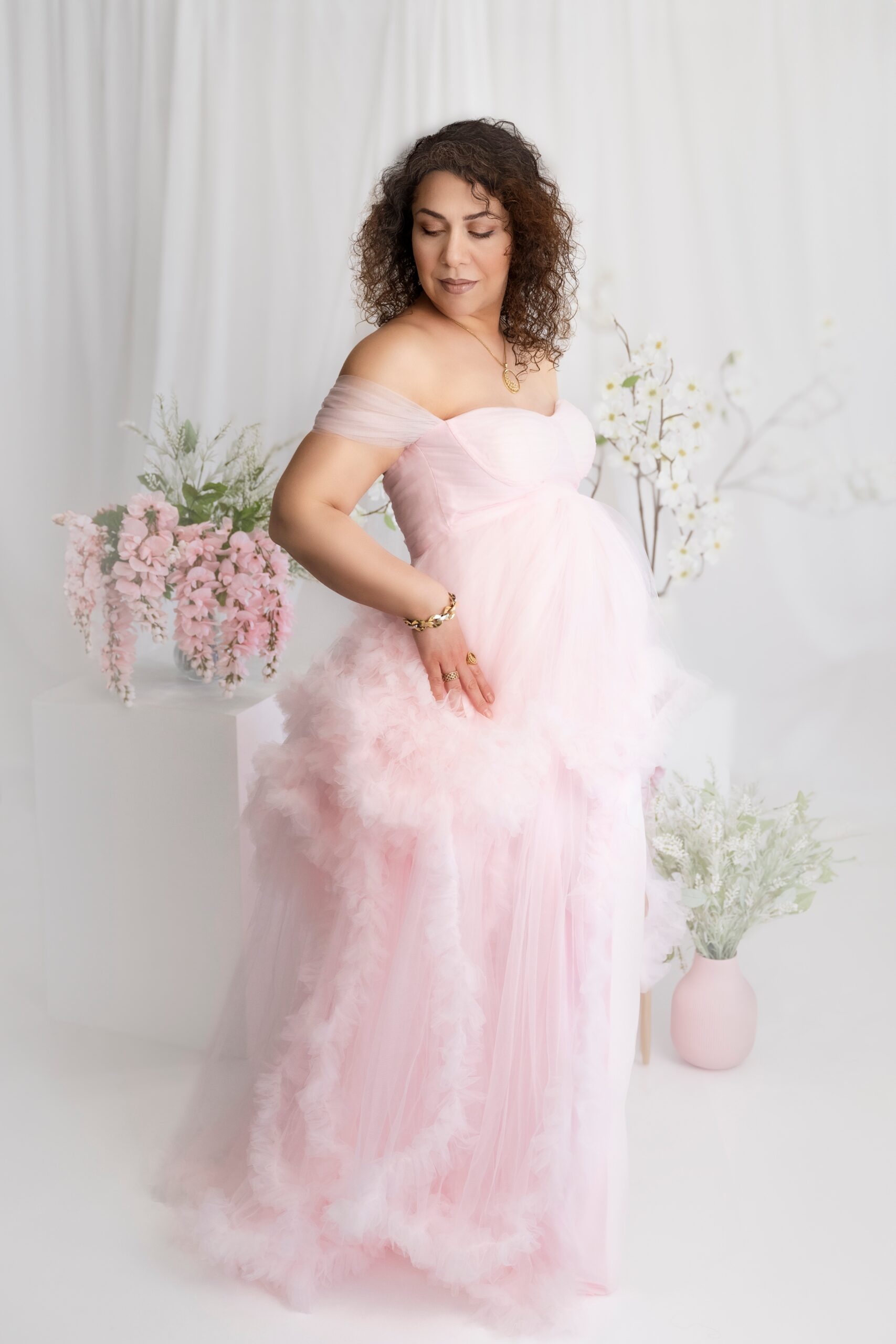 A pregnant woman stands in a pink gown in a studio looking down her shoulder after finding perfect maternity clothes in Edmonton