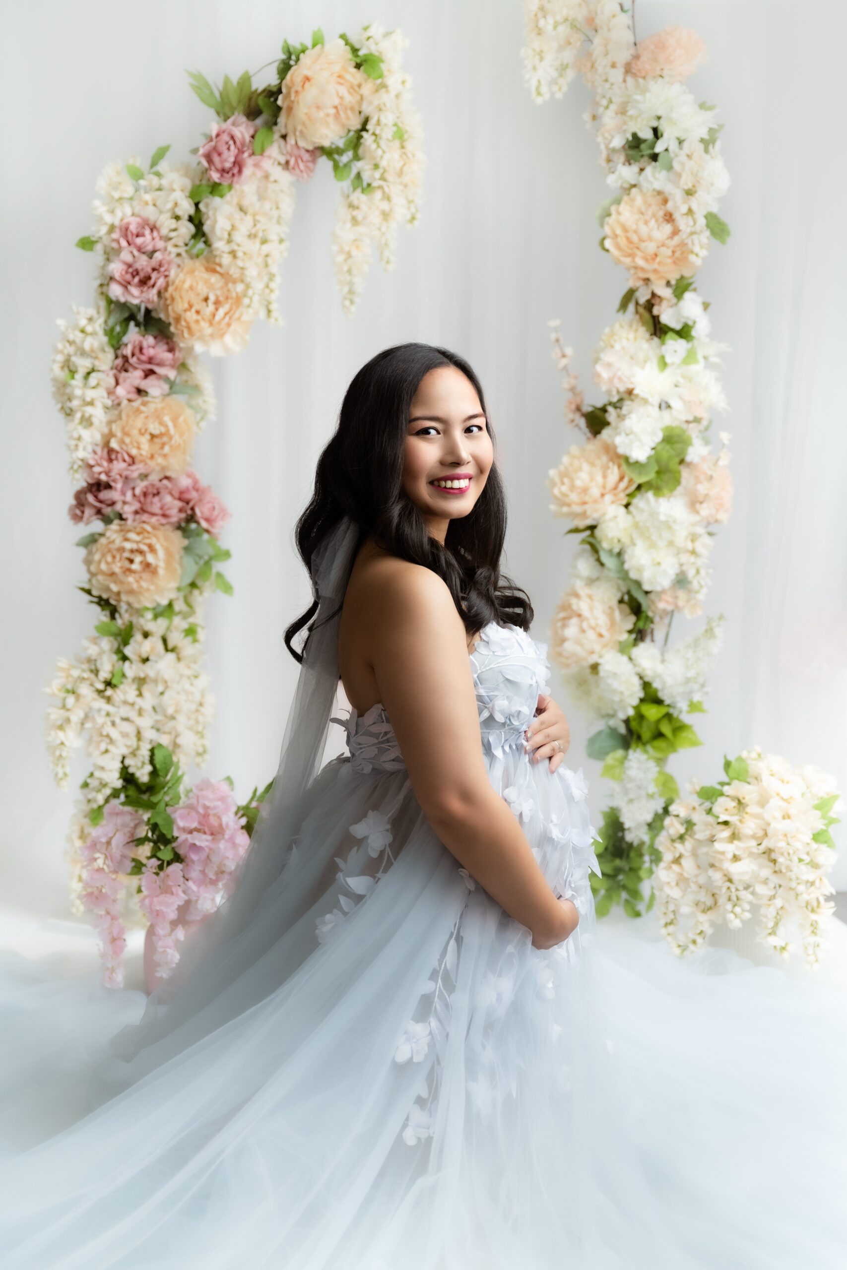 A mom to be smiles over her shoulder while standing in a studio in a long blue flowing maternity gown after shopping for maternity clothes in Edmonton