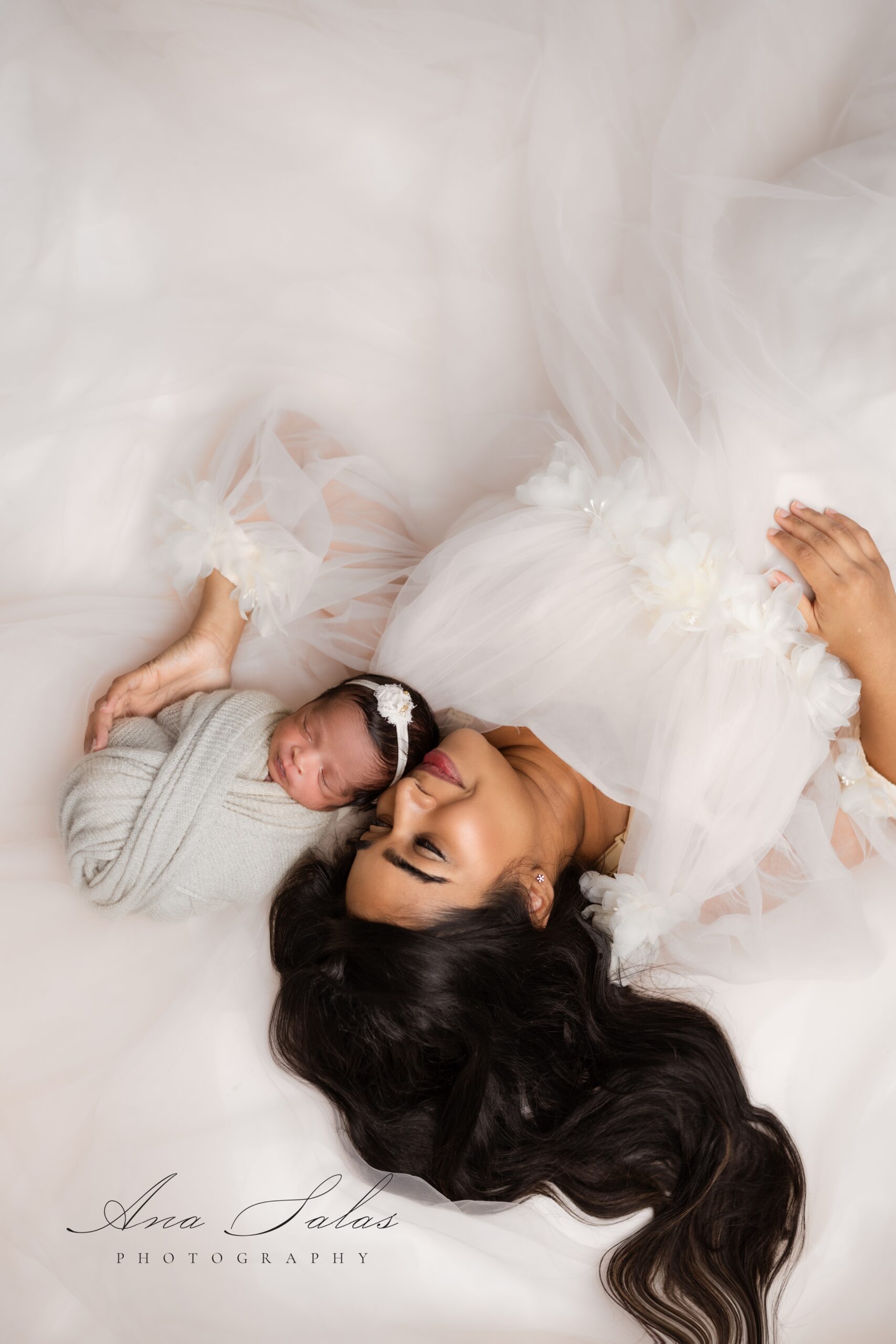 A smiling new mom in a white gown snuggles her sleeping newborn against her cheek