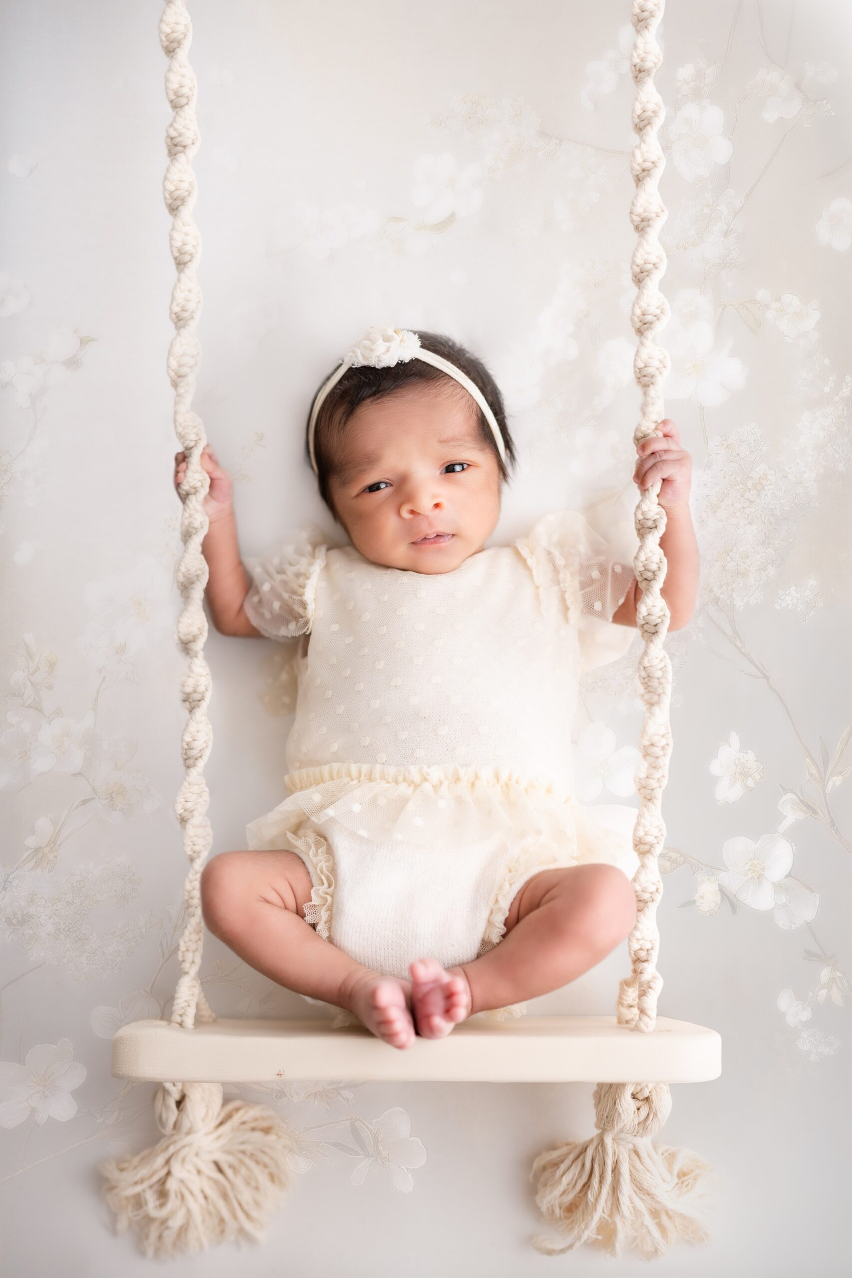 A sleepy baby sits on a swing in a white onesie and headband after visiting the fourth edmonton