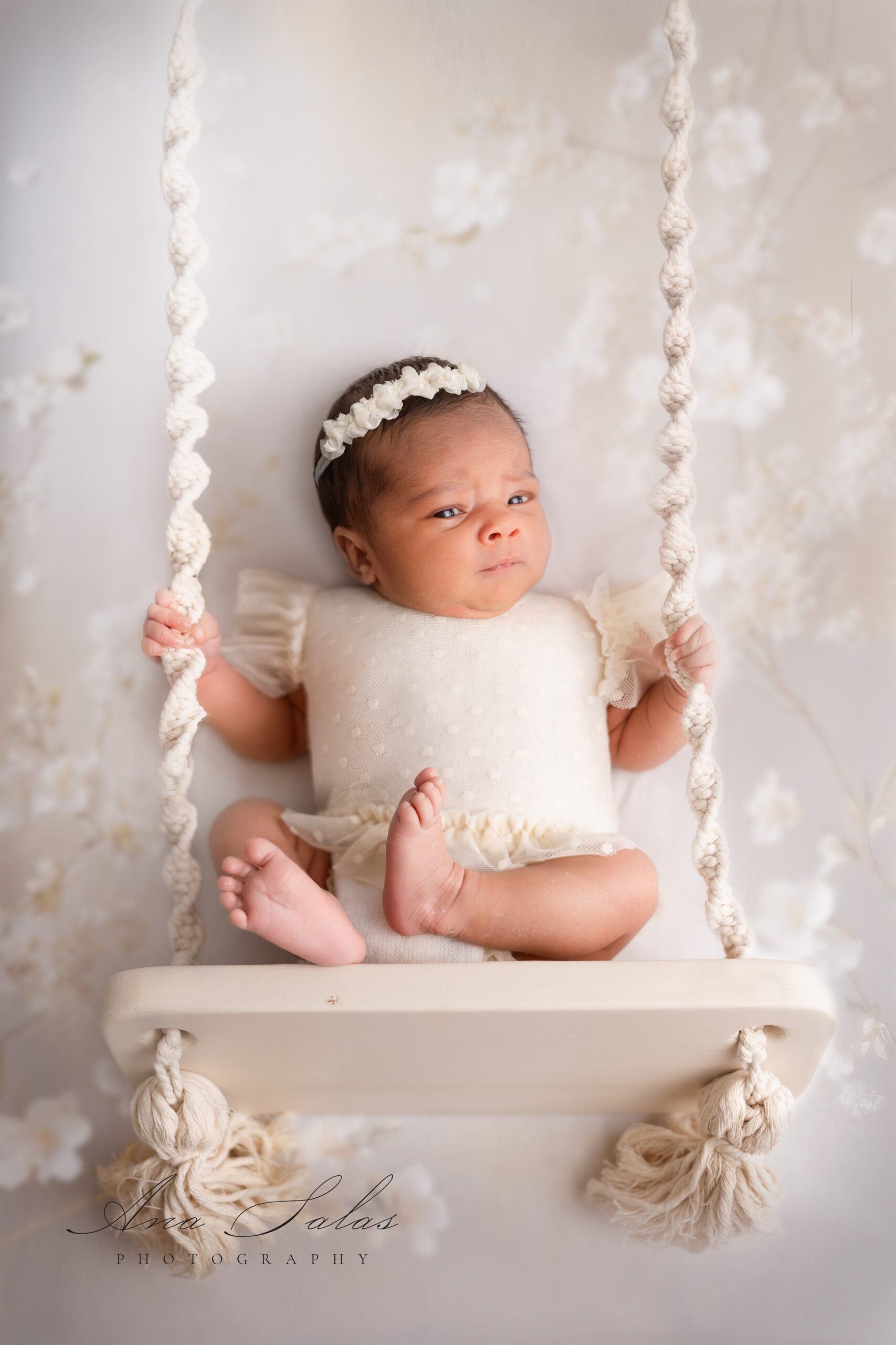 A newborn baby sits on a swing in a studio in a white dress onesie
