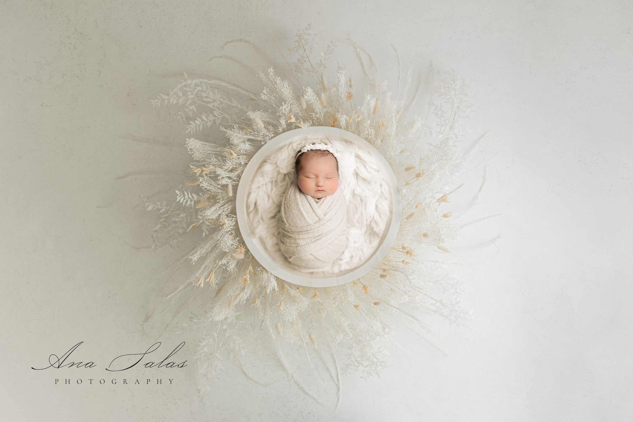 A sleeping newborn baby girl in a white bowl surrounded by matching florals in a studio before some baby classes in edmonton