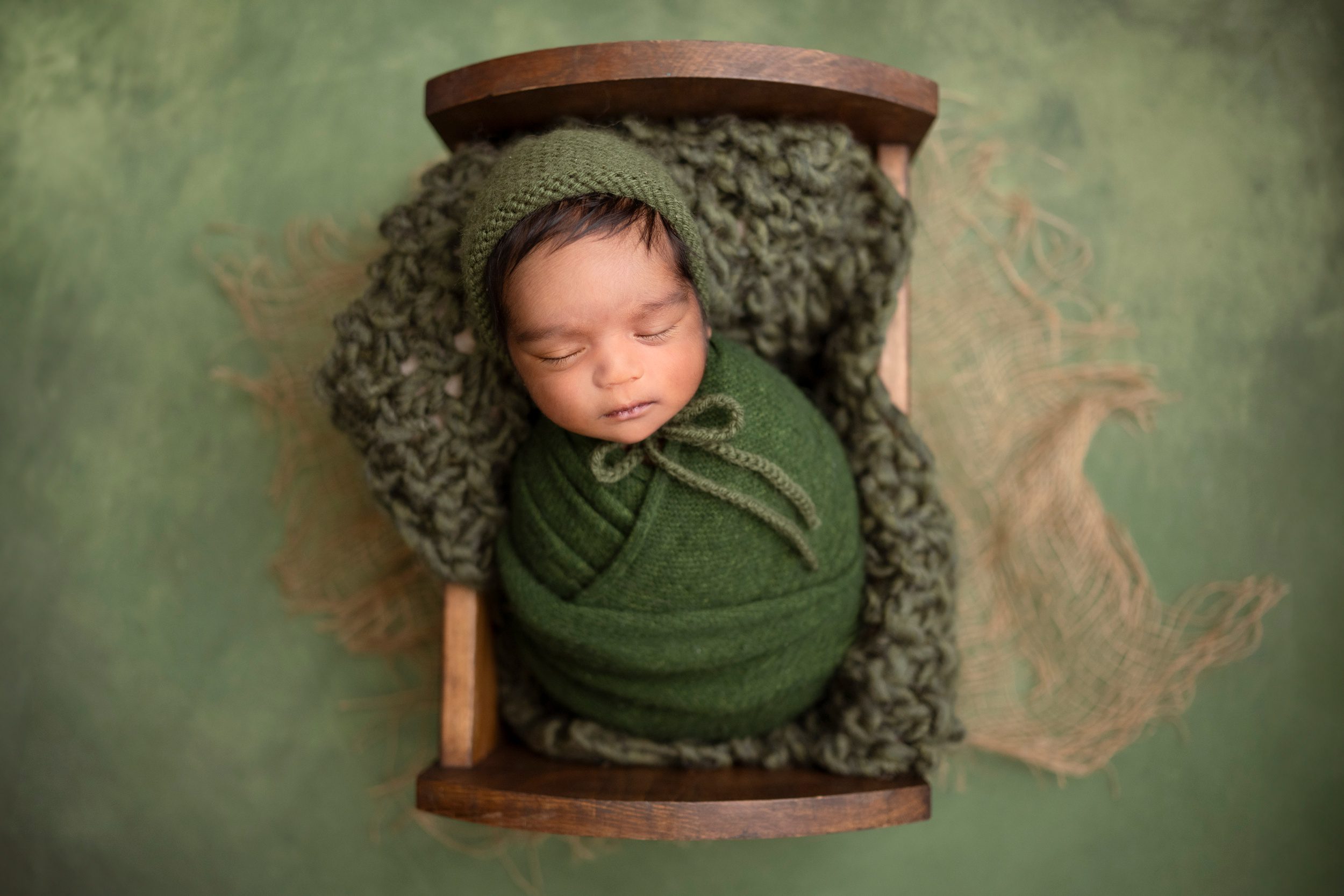 A newborn baby sleeps in a green swaddle in a tiny wooden crib in a studio